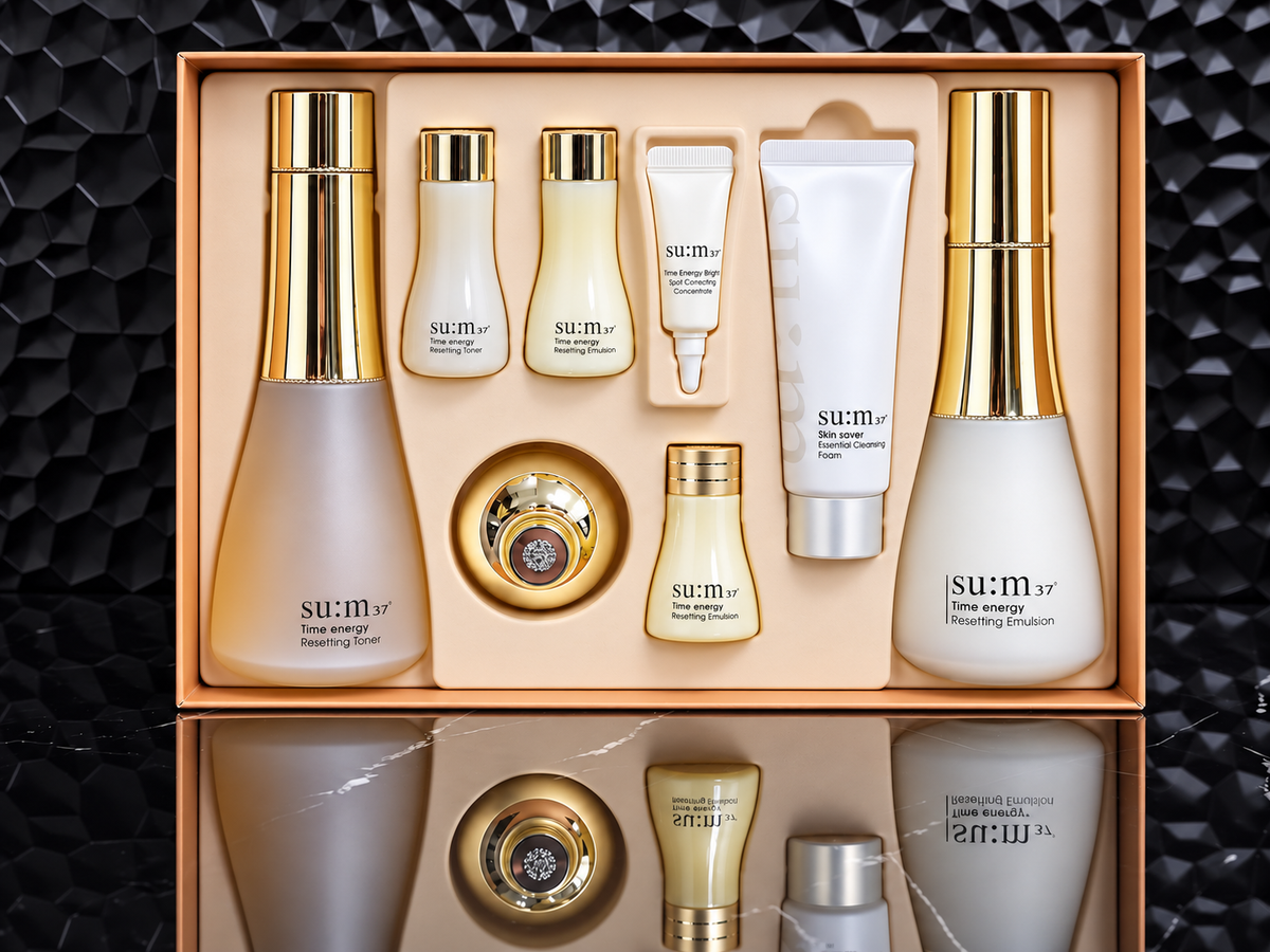 A sum:37 skincare gift set arranged in an open rectangular box, featuring multiple bottles, tubes, and a round jar with gold caps and frosted glass. The products are shown against a dark textured background with a mirrored reflection below.