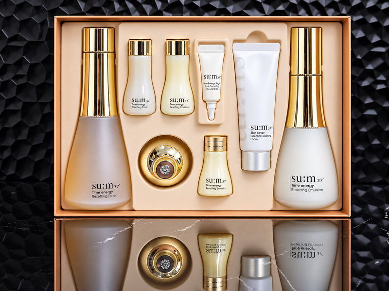 A sum:37 skincare gift set arranged in an open rectangular box, featuring multiple bottles, tubes, and a round jar with gold caps and frosted glass. The products are shown against a dark textured background with a mirrored reflection below.