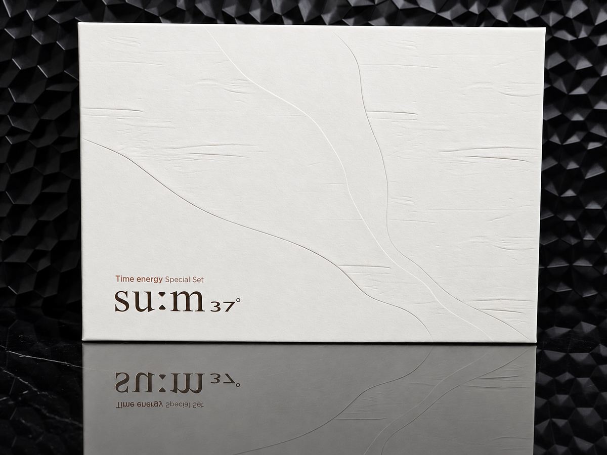 A square white product box with embossed abstract line art and the visible brand text 'su:m 37' on the lower left, photographed against a dark textured background with a reflective surface below.